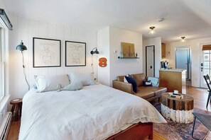 A bedroom with a bed, bedside lamps, a sofa, a coffee table, and a view of the kitchen. - Cozy condo in great location with deck, board games, & AC (Provincetown)