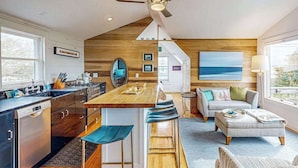 Une cuisine moderne avec un îlot central en bois, des tabourets de bar, un coin repas avec un canapé et un pouf, et une vue sur l’océan grâce à de grandes fenêtres.