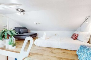 A modern loft bedroom with a sloped ceiling, a bed with white bedding, a dark sofa, a white chair, and a small table with plants. - Loft-style condo with private deck - near Truro Beach/Dune Shacks (Provincetown)