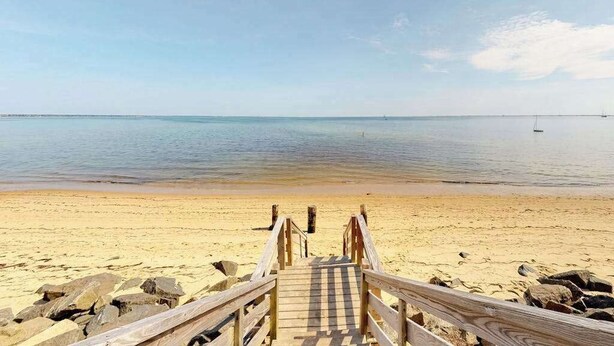 Eine Holzstege führt zu einem Sandstrand mit ruhigem Wasser und blauem Himmel.