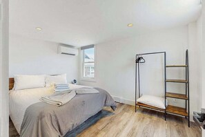 A bedroom with a bed, a window, a wall-mounted air conditioner, and a metal shelf unit. - Second-floor condo steps from downtown with large deck, partial bay view & AC (Provincetown)