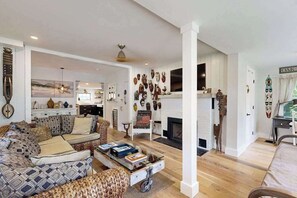 Ein Wohnzimmer mit Sofa, Couchtisch und Kamin. Es gibt eine Essecke mit Tisch und Stühlen, und im Hintergrund eine Küche.