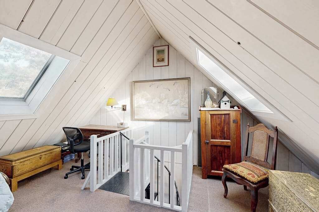 A loft bedroom with a sloped ceiling, a desk with a chair, a wooden cabinet, a chair with a patterned cushion, and a framed picture on the wall.