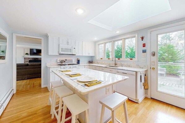 A modern kitchen with white cabinetry, a central island with bar stools, a built-in microwave, and a large window overlooking greenery. - Tranquil & sunlit home with peaceful yard, large deck, & fireplace - near beach (Orleans)