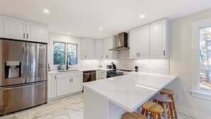 Una cucina moderna con mobili bianchi, un frigorifero in acciaio inossidabile, una grande isola con sgabelli e una finestra che lascia entrare luce naturale.