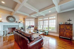 A spacious living room with a fireplace, leather sofa, wooden flooring, and large windows.