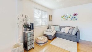 A living room with a grey sofa, a white rug, a side table with a vase of flowers, and a window with blinds. - Lakefront home with private dock, deck, & lovely view - short drive to the cape (Plymouth)