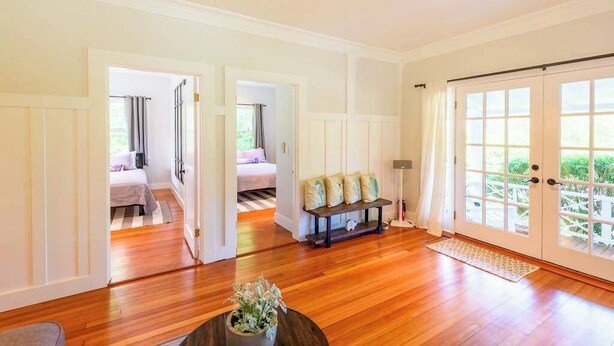Quarto espaçoso com piso de madeira, um banco com almofadas, uma porta que dá para uma varanda e uma planta em vaso sobre uma mesa.