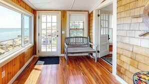 A wooden interior with a bench, a door leading outside, and a view of the ocean. - Lovely oceanfront cottage with deck, grill, & panoramic views (Plymouth)