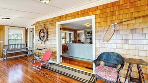 A room with wooden panel walls, a wooden floor, a bench, a red chair, a wicker chair, and a small table. - Lovely oceanfront cottage with deck, grill, & panoramic views (Plymouth)