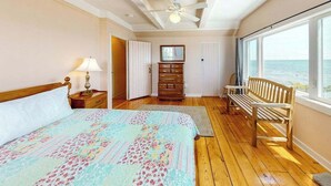 A bedroom with a bed, a wooden bench, a dresser, a ceiling fan, and a view of the sea. - Lovely oceanfront cottage with deck, grill, & panoramic views (Plymouth)