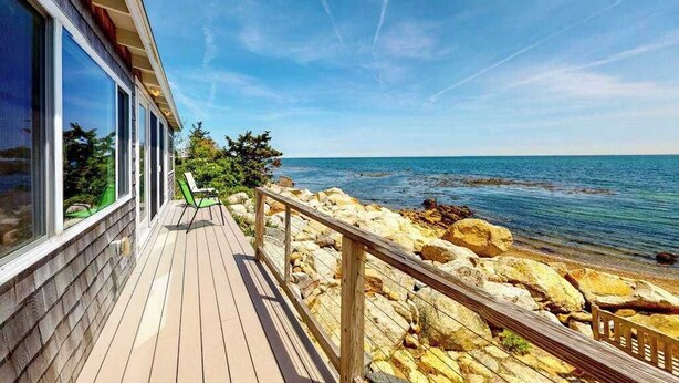 A wooden deck with a railing, overlooking a rocky shoreline and the sea. - Lovely oceanfront cottage with deck, grill, & panoramic views (Plymouth)