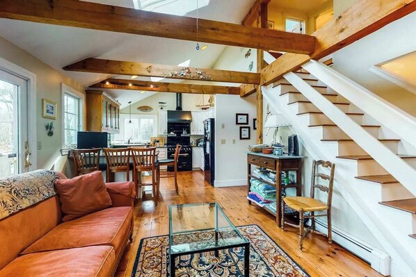 A spacious living area with a wooden staircase, a sofa, a glass coffee table, and a kitchen in the background.
