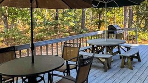 An outdoor deck with a round table and chairs, a grill, and a view of a wooded area. - Quiet home with sunroom, deck, W/D, AC, & quick access to beach (Wellfleet)