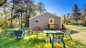 Ett litet hus med en veranda, ett bord och stolar på en gräsig gård omgiven av träd.