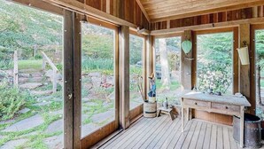 A wooden room with large windows, a wooden bench, and a rustic outdoor view. - Nana’s Treehouse | House with spacious deck, grills, outdoor shower & W/D (West Tisbury)