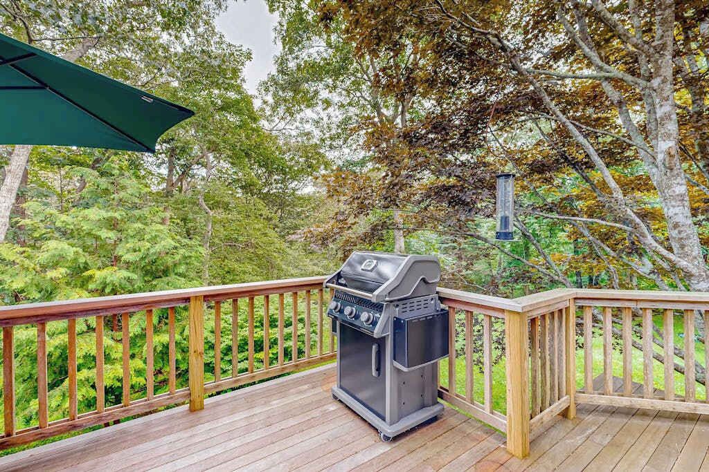 A barbecue grill on a wooden deck with a railing, surrounded by trees.