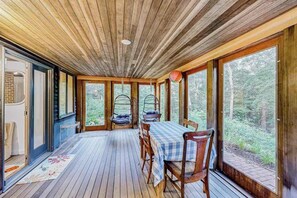 A wooden deck with a table and chairs, large windows, and hanging chairs. - Nana’s Treehouse | House with spacious deck, grills, outdoor shower & W/D (West Tisbury)