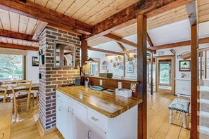 A kitchen with a brick wall, wooden beams, a wooden countertop, and a dining area with chairs and a table. - Nana’s Treehouse | House with spacious deck, grills, outdoor shower & W/D (West Tisbury)