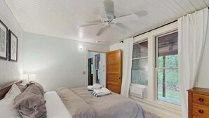Un dormitorio con cama, ventilador de techo, ventana con cortinas, puerta de madera y cómoda.