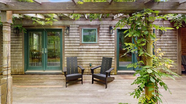 Una terraza de madera con dos sillas Adirondack, un pérgola y una casa de madera con puertas y ventanas verdes.
