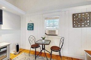 Une pièce aux murs recouverts de panneaux blancs, une petite table avec deux chaises, une enseigne suspendue, une fenêtre avec des stores et un élément décoratif mural.