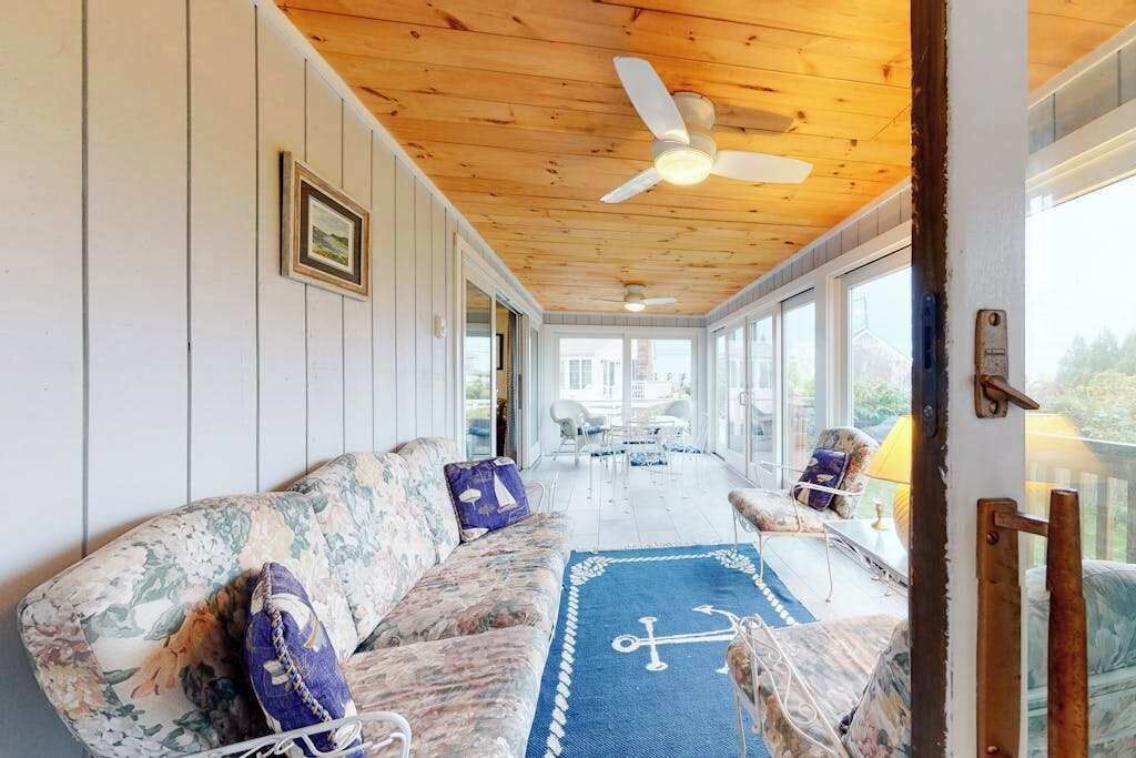 A sunlit room with a wooden ceiling, a sofa, a coffee table, and a view through large windows.