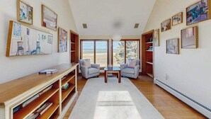 Um quarto espaçoso com uma grande janela, uma escrivaninha de madeira, duas poltronas e uma estante de livros.