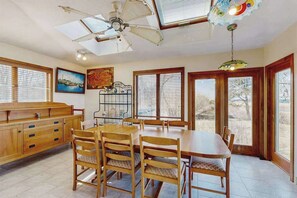 Un comedor con una mesa y sillas de madera, un ventilador de techo y ventanas grandes con persianas.