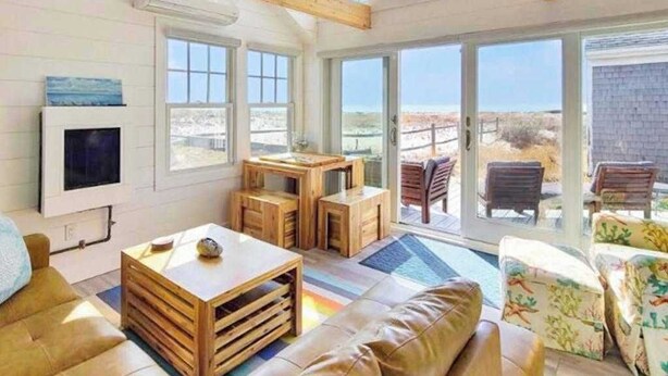 A spacious room with a large window, a wooden ceiling, a sofa, a coffee table, and a television. - Peaceful Beachfront Cottage with Bay Views: near town, bike trails & dune walks (North Truro)