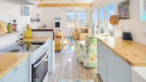 A kitchen with wooden beams, a stove, and a dining area with a colorful rug. - Peaceful Beachfront Cottage with Bay Views: near town, bike trails & dune walks (North Truro)