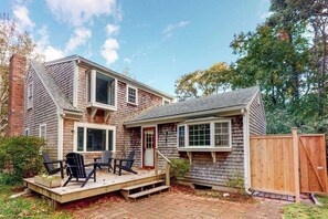 A two-story house with a deck, outdoor seating, and a wooden fence. - Peaceful half-acre home near ocean, dining & shops and a fire pit (Wellfleet)