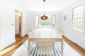 A spacious room with a long white table, white chairs, a woven pendant light, and large windows. - Peaceful half-acre home near ocean, dining & shops and a fire pit (Wellfleet)