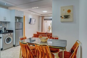 A modern kitchen with a washing machine, a dining area with a glass table and orange chairs, and a living area with a sofa and TV. - Studio with Terrace Overlooking the Beach (Tamraght)