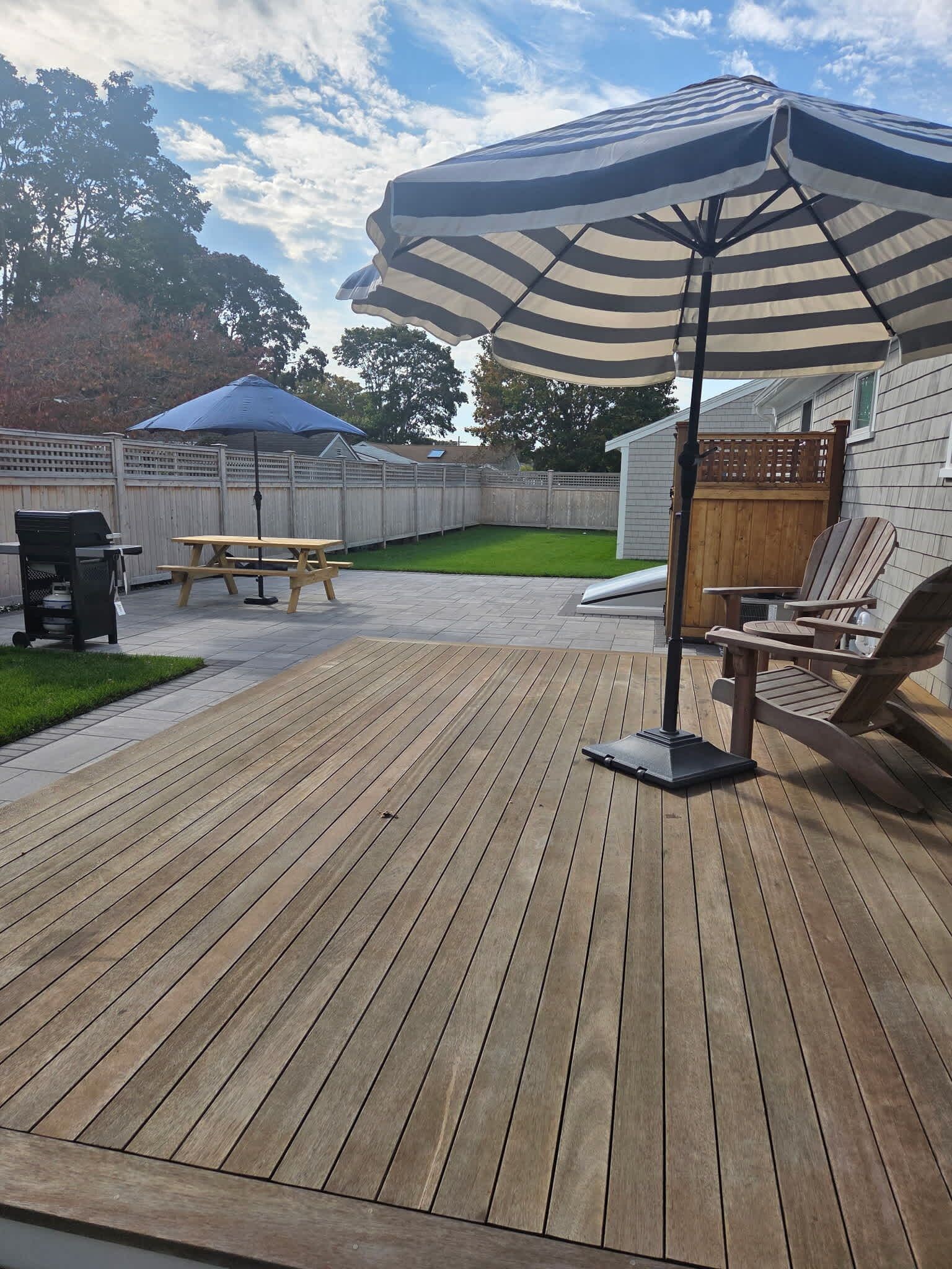 A wooden deck with a picnic table and a grill, a large striped umbrella, and a wooden fence.