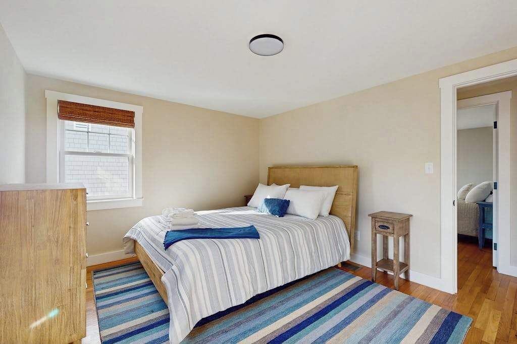 A bedroom with a bed, a wooden dresser, a window with blinds, a bedside table, and a door leading to another room.