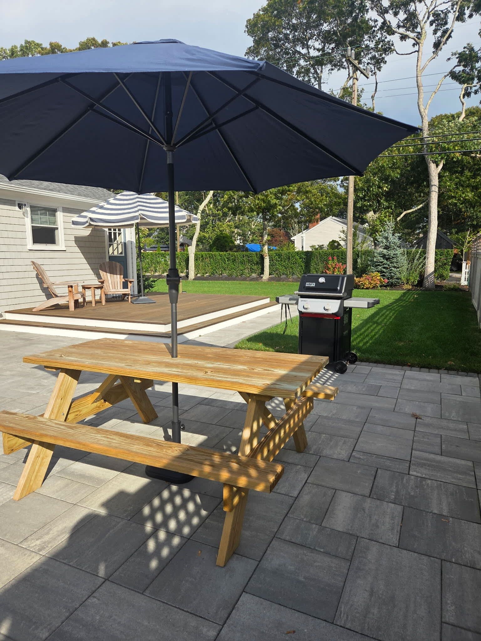 A wooden picnic table with an umbrella, a barbecue grill, and a lawn in the background.