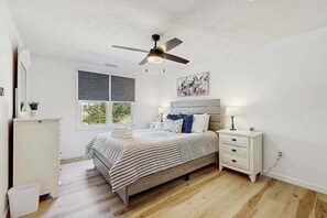 Ein Schlafzimmer mit Bett, Nachttisch, Deckenventilator, Fenster und einem Wandbild.