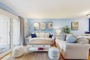 A living room with a sofa, coffee table, and two poufs, a lamp, and wall art. - Modern fairway condo with open layout, private patio, AC & washer/dryer (Brewster)