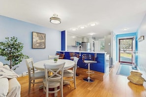 A compact kitchen with modern appliances, a dining area with white chairs, and a blue accent wall. - Modern fairway condo with open layout, private patio, AC & washer/dryer (Brewster)