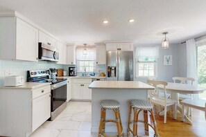 Una cocina moderna con muebles blancos, una isla central, taburetes de bar y un comedor con sillas blancas.