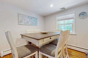 A dining area with a wooden table, white chairs, a map on the wall, a window with blinds, and a clock.