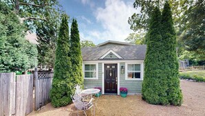 Een gelijkvloers huis met een voordeur, een klein terras met een tafel en stoelen, en twee grote altijdgroene bomen.