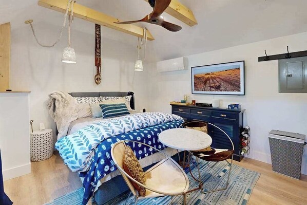 Une chambre à coucher avec un lit, une table ronde, une chaise, un ventilateur de plafond, un téléviseur fixé au mur et un tableau encadré.