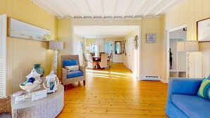 Ein Zimmer mit Holzboden, einem blauen Sofa, Korbsesseln, einem Esstisch und einer weißen Decke.