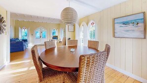 Ein Essbereich mit einem runden Tisch, Korbstühlen, einer Chandelierleuchte und einem Gemälde mit Strandmotiv an der Wand.
