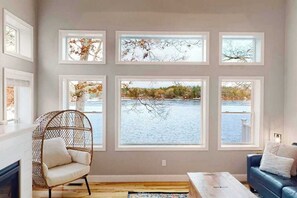 Ein Zimmer mit großem Fenster mit Blick auf ein Gewässer, ein Korbsessel, ein blaues Sofa und ein Holztischchen.