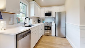 A modern kitchen with white cabinets, stainless steel appliances, a dishwasher, and a window. - Beautifully remodeled cottage near the beach with central AC, gas grill & lawn (Orleans)