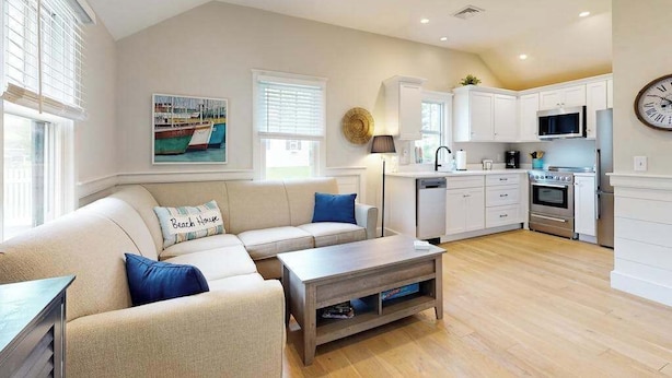 A modern living room with a sofa, coffee table, and a kitchen area with cabinets and appliances. - Beautifully remodeled cottage near the beach with central AC, gas grill & lawn (Orleans)