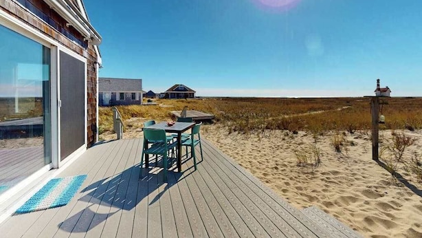 A wooden deck with outdoor furniture, a glass door entrance, and a view of the beach and dunes. - Cozy cottage steps to the beach with patio, W/D, & partial AC (North Truro)